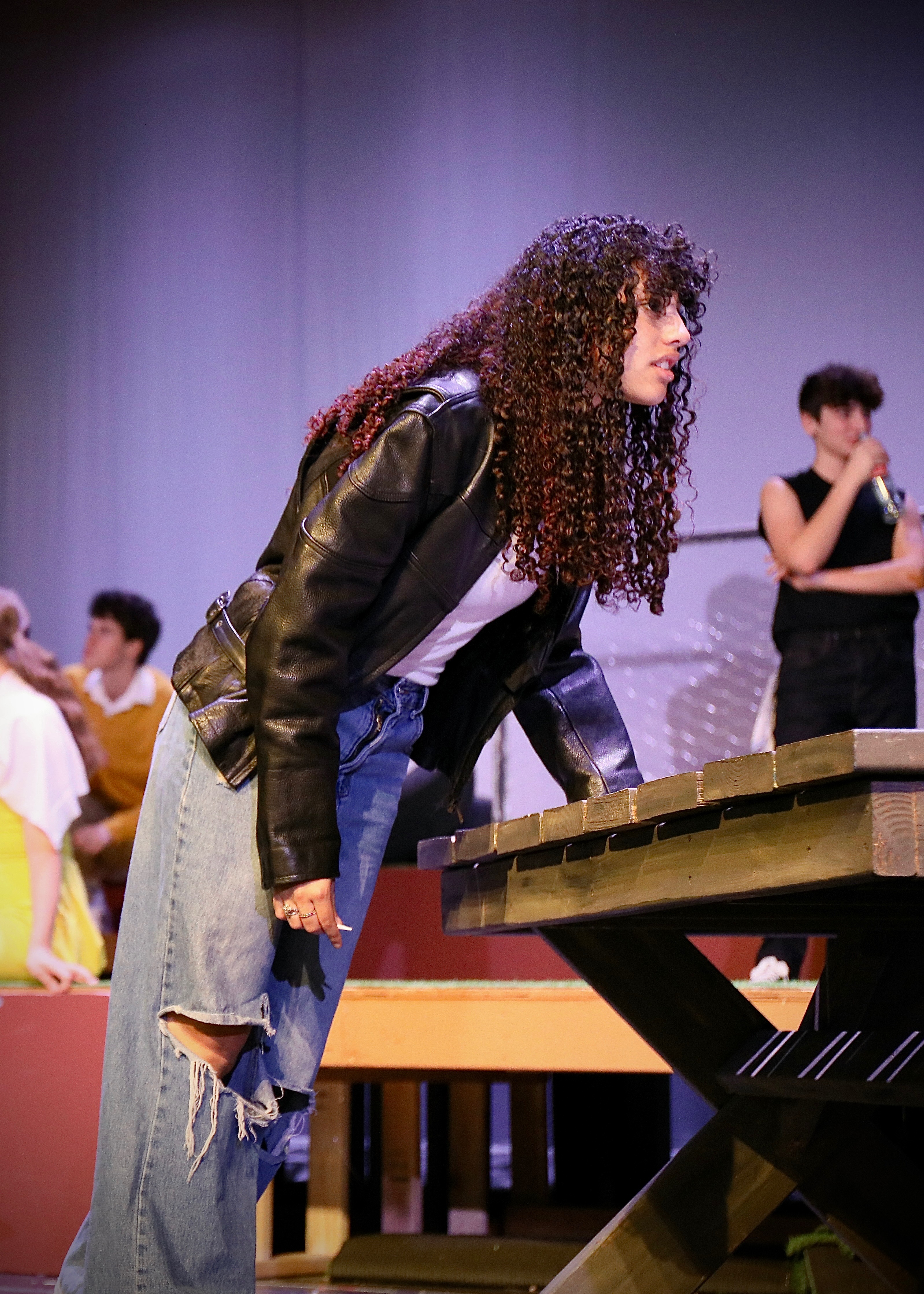 Actor in leather jacket leans over a picnic table on stage